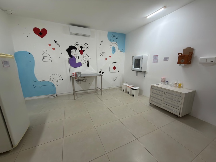 Sala de quimioterapia da COEV, com mesa de aplicação em inox, descarte segregado e mural ilustrado de tutora abraçando um cão